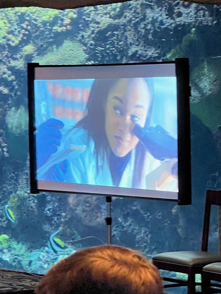 A screen showing a woman in a lab coat and gloves examining something. Behind, an aquarium.