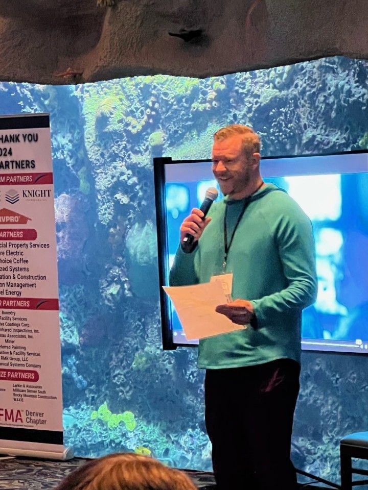 Man speaking into a microphone, smiling, holding papers. Standing in front of an aquarium tank and screen.