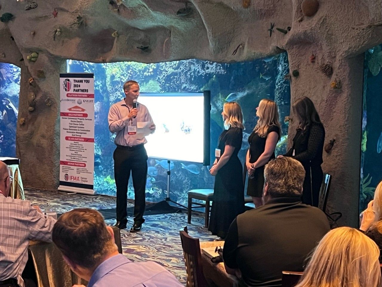 A man presents at a podium. Three women stand nearby. A presentation screen is behind them.