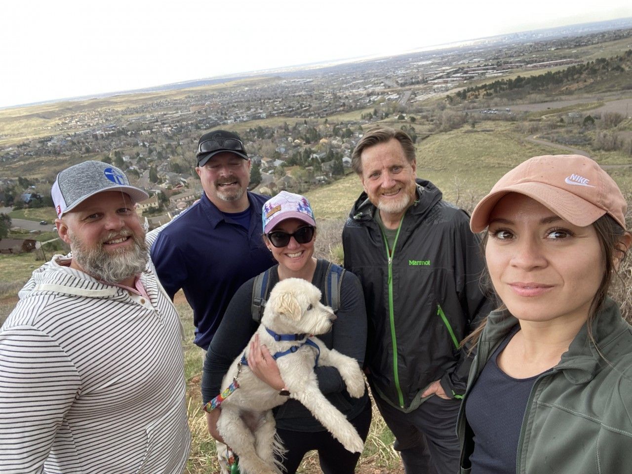 Group of people with a dog on a hillside overlooking a town. Some wear hats and casual clothes. Cloudy sky.