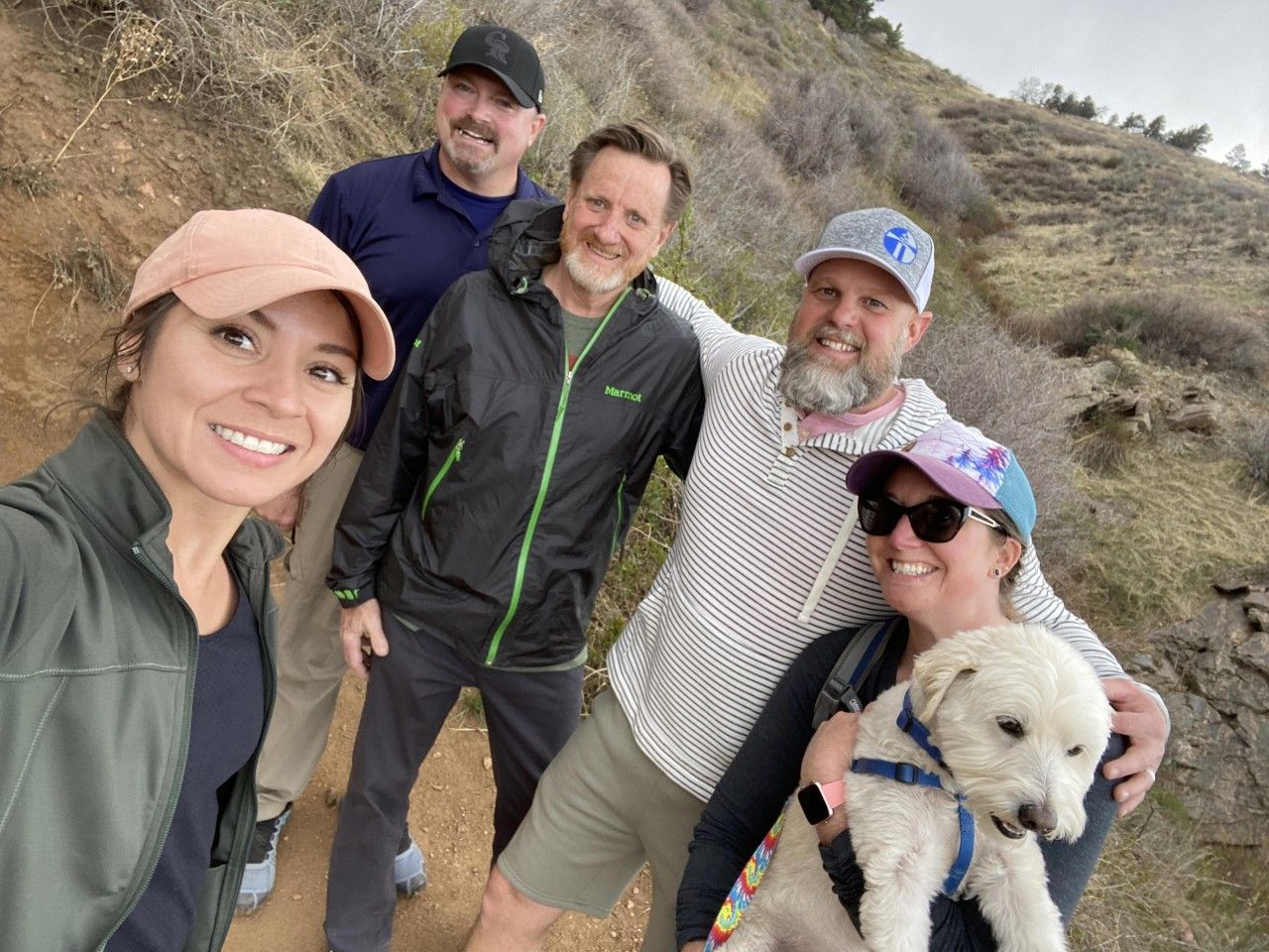 Group of five people hiking on a trail. One person holding a white dog. All smiling at the camera.