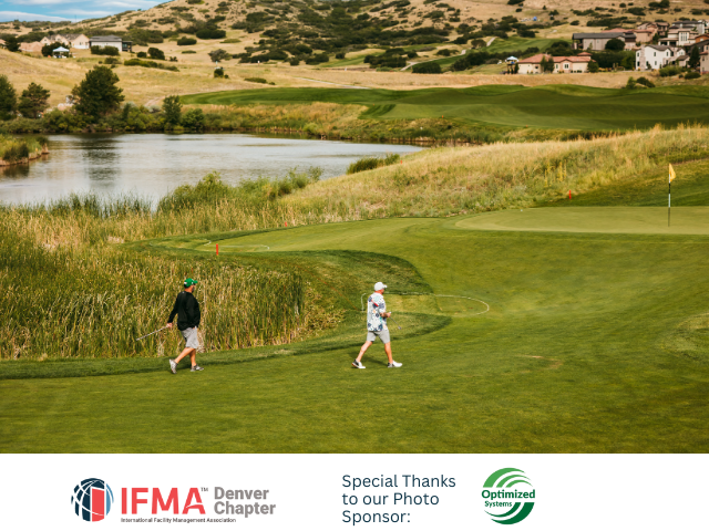 Two golfers walking on a green golf course, a lake, and hills in the background.
