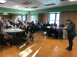Woman speaking at a presentation in a room; audience seated at round tables.