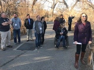Group of people on a paved path outdoors. One person in a wheelchair. Trees in the background, sunny.