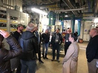 Group of people touring a utility room with industrial equipment, pipes, and electrical panels.