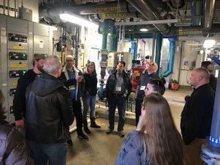 A group of people in an industrial setting, possibly on a tour. Electrical panels and pipes are visible.