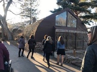People walking towards a brown arched building with large windows. Sunshine casts shadows.