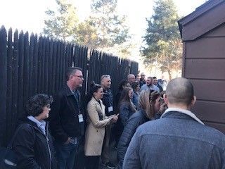 Group of people gathered outside, near a dark wooden fence and building, outdoors on a sunny day.