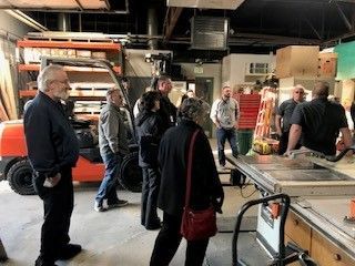 People gathered in a workshop with machinery, including a forklift and a saw.