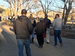 People walking along a path on a sunny day. Trees and buildings are in the background.