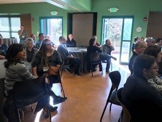 People seated, listening in a room. Some face the camera; others face a table with people. Bright room with doors.