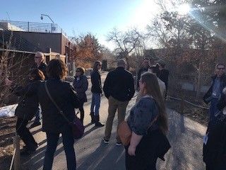 A group of people standing outside on a sunny day. They appear to be on a tour, looking at a building.
