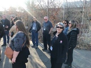 A group of people standing outside, some wearing sunglasses, near a fence, on a sunny day.