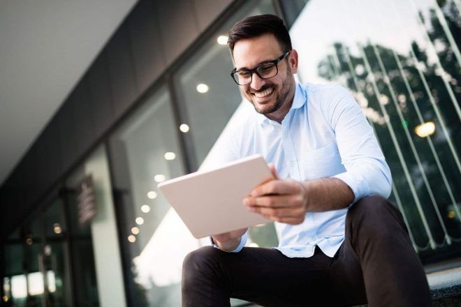 Man in glasses smiles while using tablet outdoors.