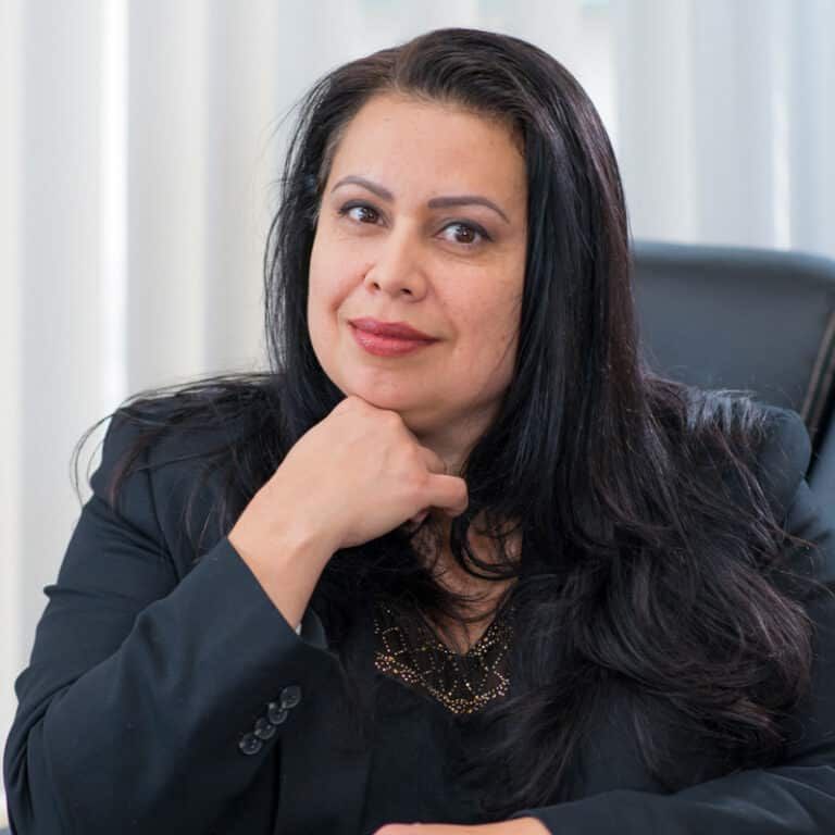 Woman with dark hair, in a blazer, rests chin on hand, smiling slightly, in an office setting.