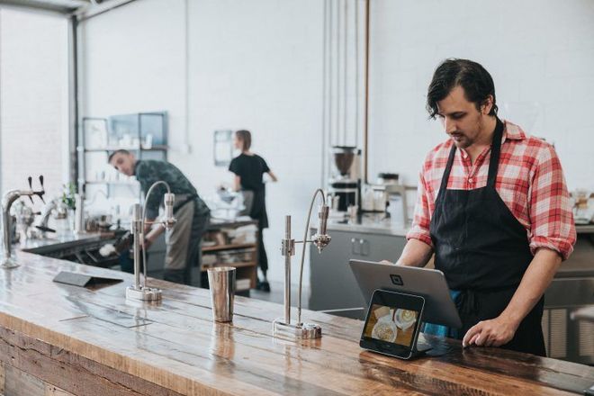 Man in red plaid shirt and black apron uses a tablet at a cafe counter; two other employees in the background.