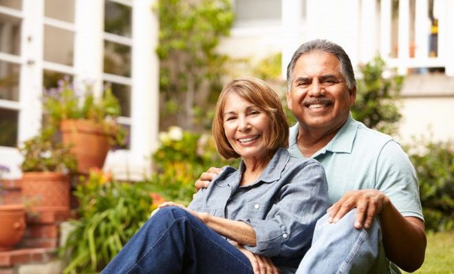 Smiling senior couple in garden, woman seated with man's arm around her, house in background.