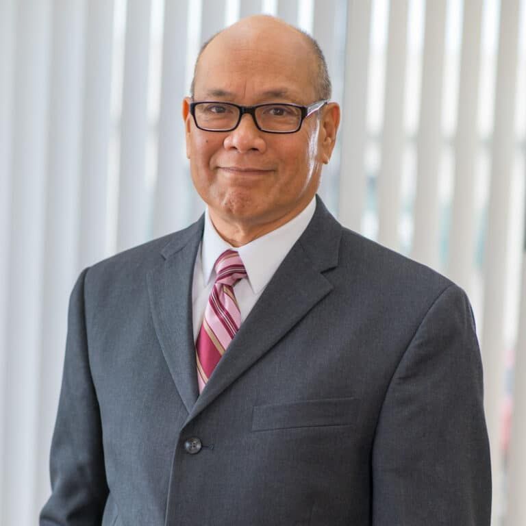 Man in a suit, wearing glasses, smiles. Indoors, near a window.