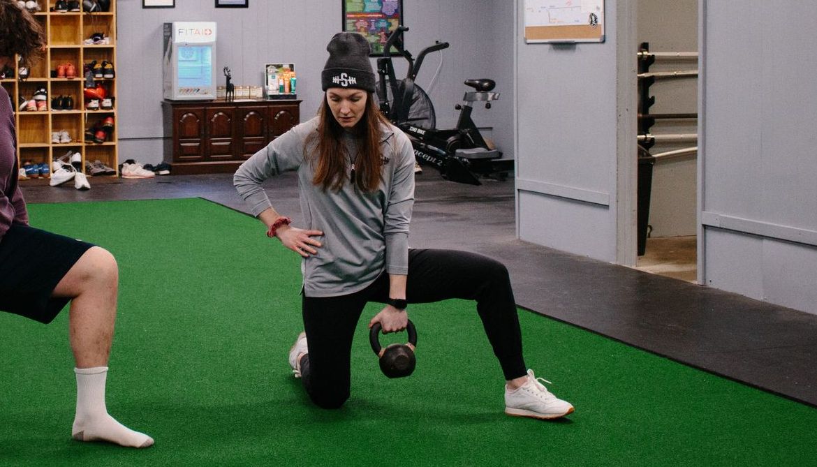 A woman is kneeling down while holding a kettlebell in a gym.