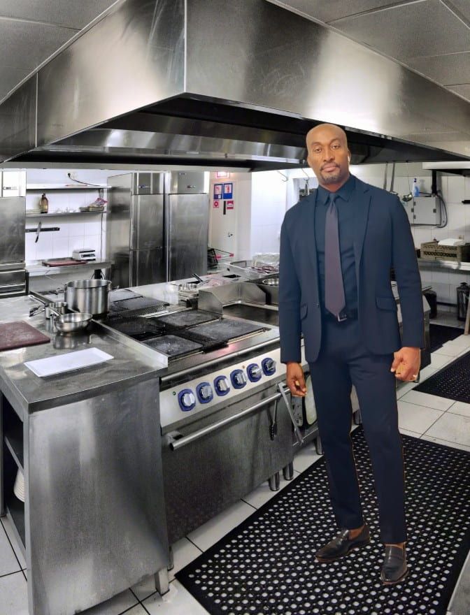 Man in a suit stands in a commercial kitchen with stainless steel appliances.