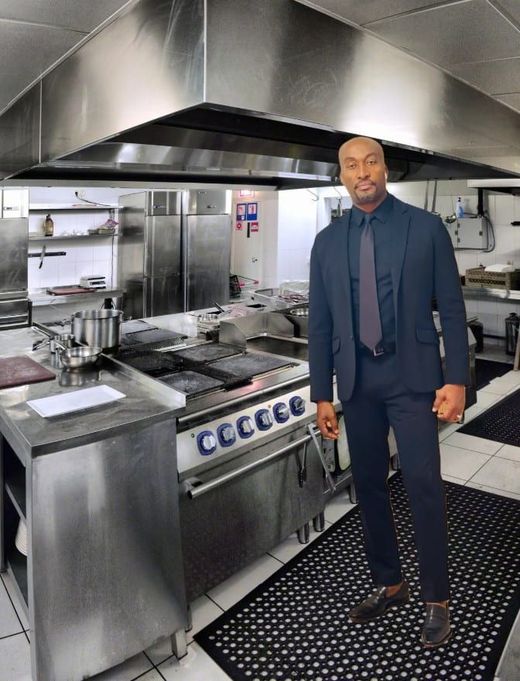 Man in a suit stands in a commercial kitchen with stainless steel appliances.
