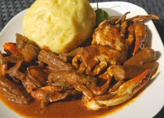 A plate with a large, golden, round dumpling served alongside a cooked crab in a rich, dark brown, okra-based sauce.