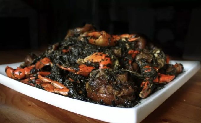 A square white platter piled high with cooked crab pieces coated in a dark, textured green herb sauce.
