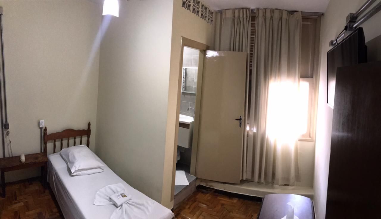 Quarto de hotel pequeno com uma cama de solteiro, porta aberta para o banheiro e janela com cortinas.