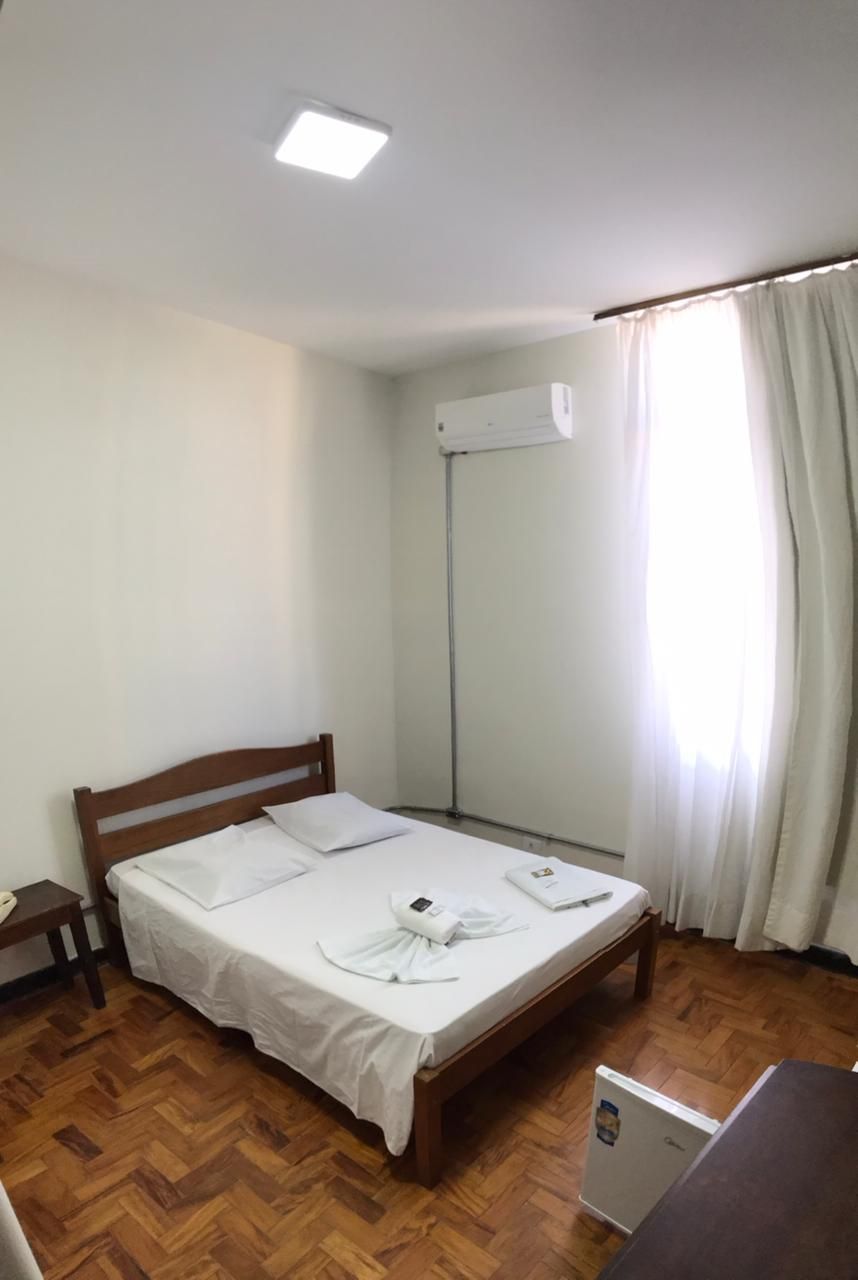 Quarto com cama de madeira, lençóis brancos, janela com cortinas translúcidas e ar condicionado.