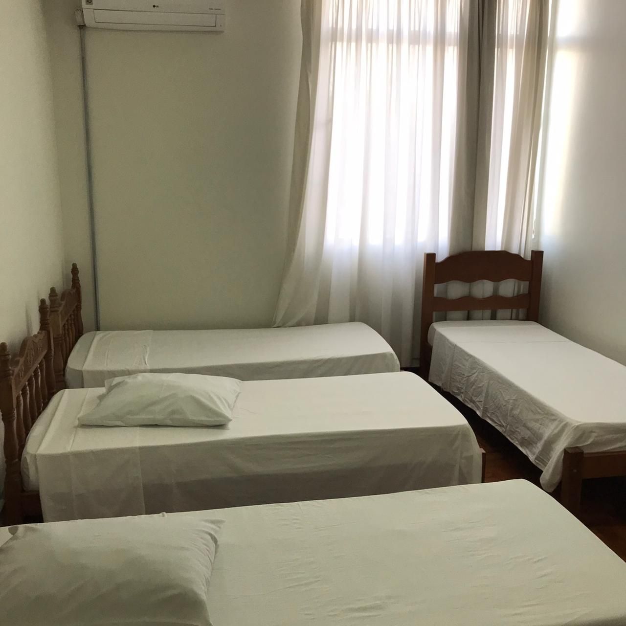 Quatro camas em um quarto, com lençóis brancos. Janela com cortinas transparentes ao fundo.