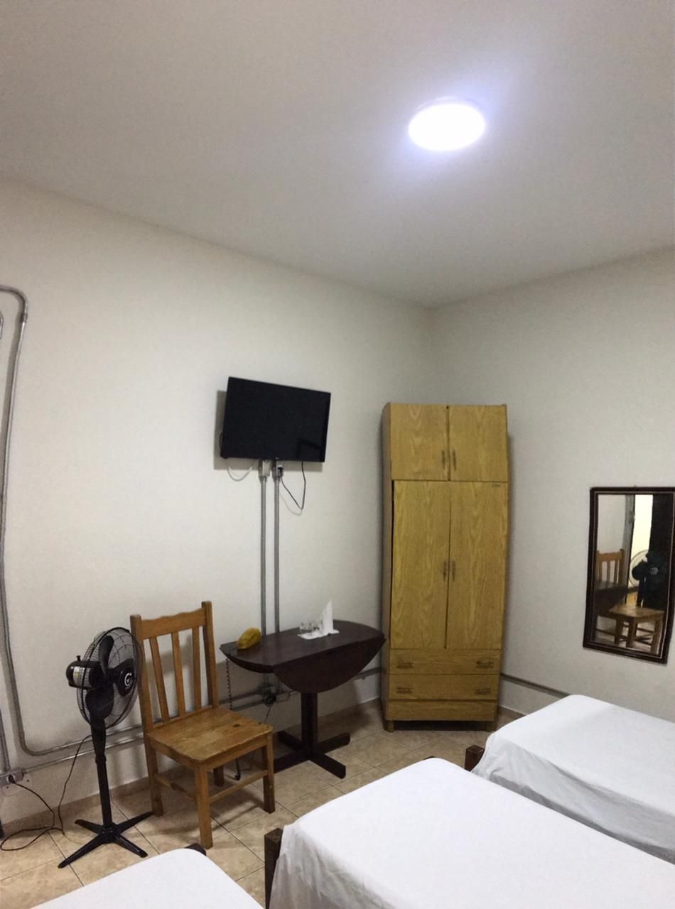 Quarto com camas, armário, TV, cadeira e mesa. Possui ventilador.