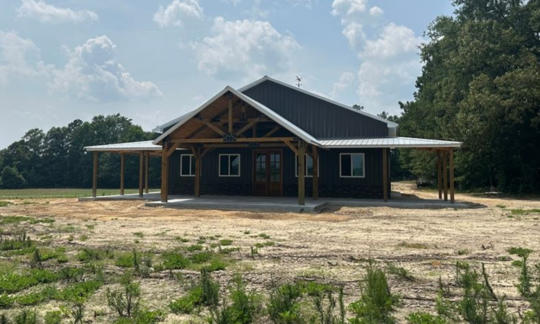 Dark gray barn-style building with a covered porch under a blue sky, set in a field.