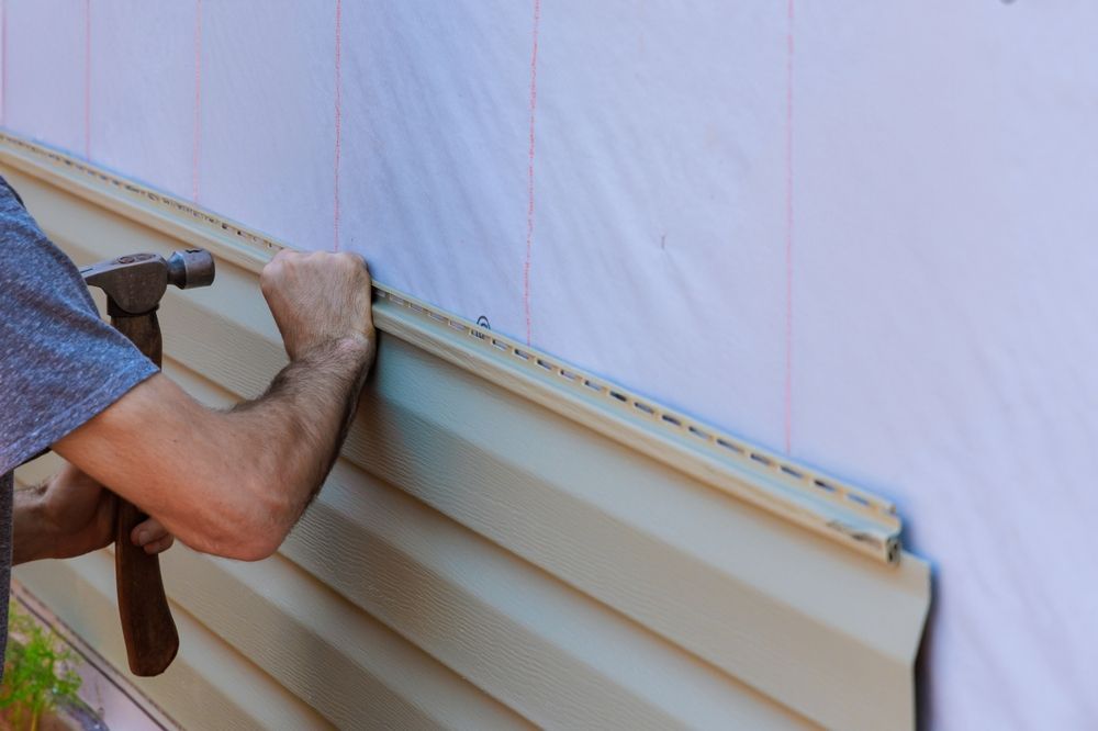 Person hammering beige vinyl siding onto a white wall.