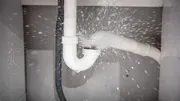 Water sprays forcefully from a disconnected joint in white PVC under-sink plumbing pipes.