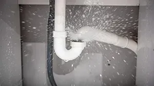 Water sprays forcefully from a disconnected joint in white PVC under-sink plumbing pipes.