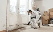 A person in protective gear, mask, and backpack sprayer treating the base of a door frame for mold or pests.