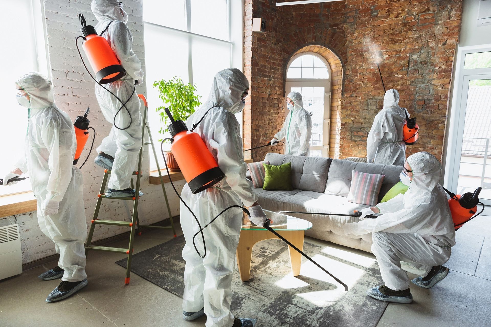 Five people in protective white hazmat suits and orange sprayers sanitize the interior of a brick-walled living room.