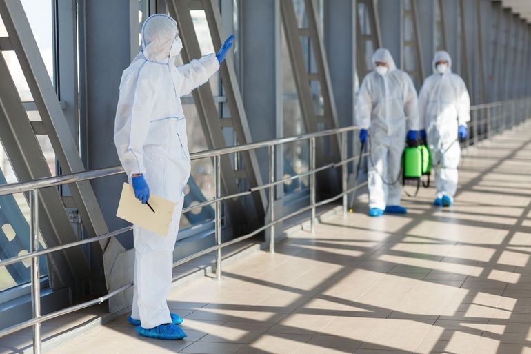 Three people in white hazmat suits and blue gloves in a hallway, with one pointing while the others carry disinfection gear.