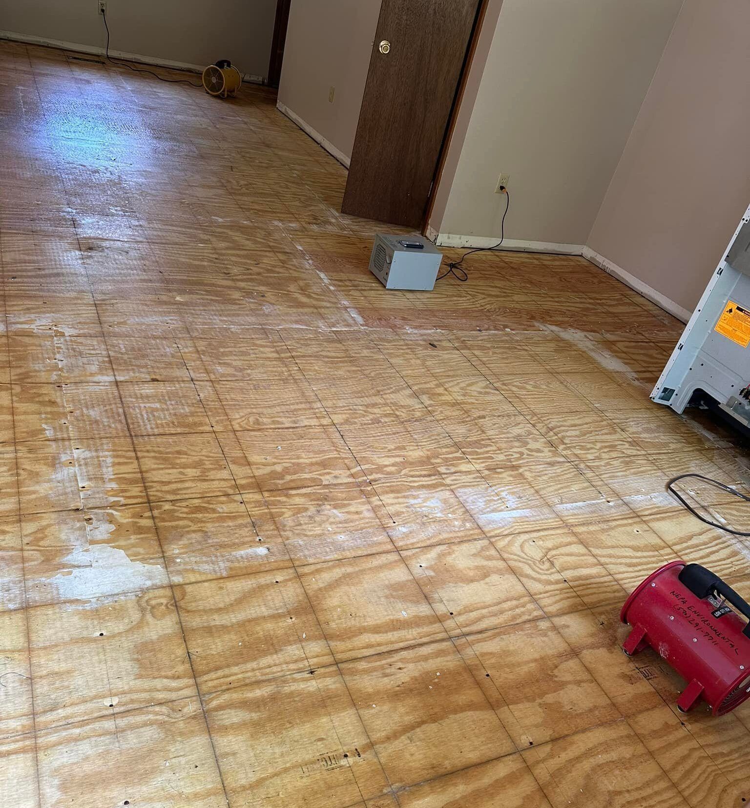 An interior room with exposed plywood subflooring, a red portable heater, and a grey air scrubber, indicating restoration.