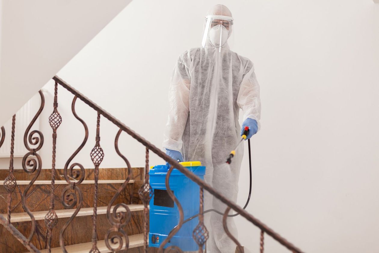 A person in full protective gear and a mask sanitizing an indoor staircase with a blue handheld sprayer.