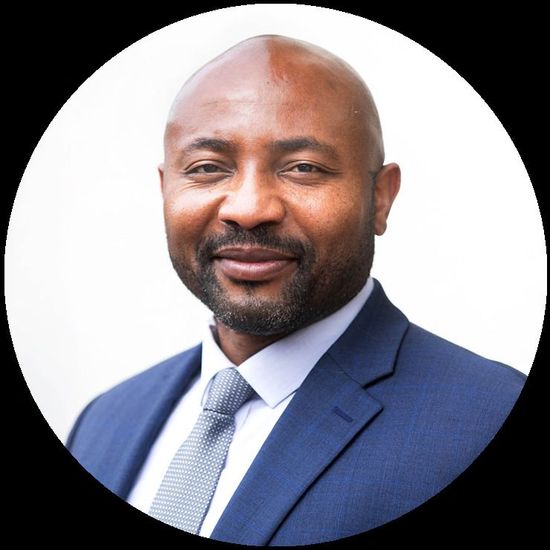 Man in a blue suit and tie, smiling in a well-lit, neutral setting.