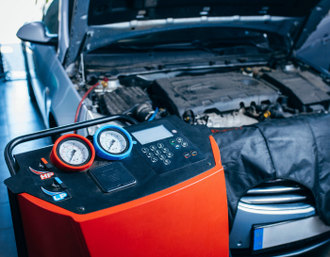 A car's engine bay is open, with an orange A/C service machine in front. Gauges and display visible | Mike's Automotive AC