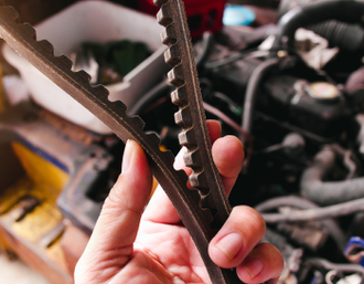 A person holds a worn, ridged engine belt in front of a blurred car engine | Mike's Automotive AC