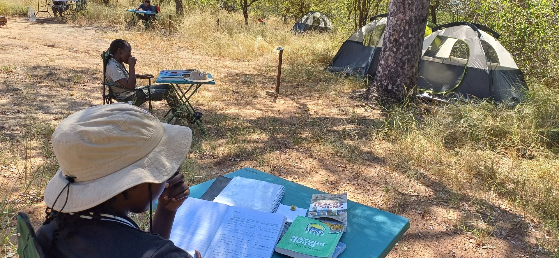 A man is sitting at a table in the woods reading a book.