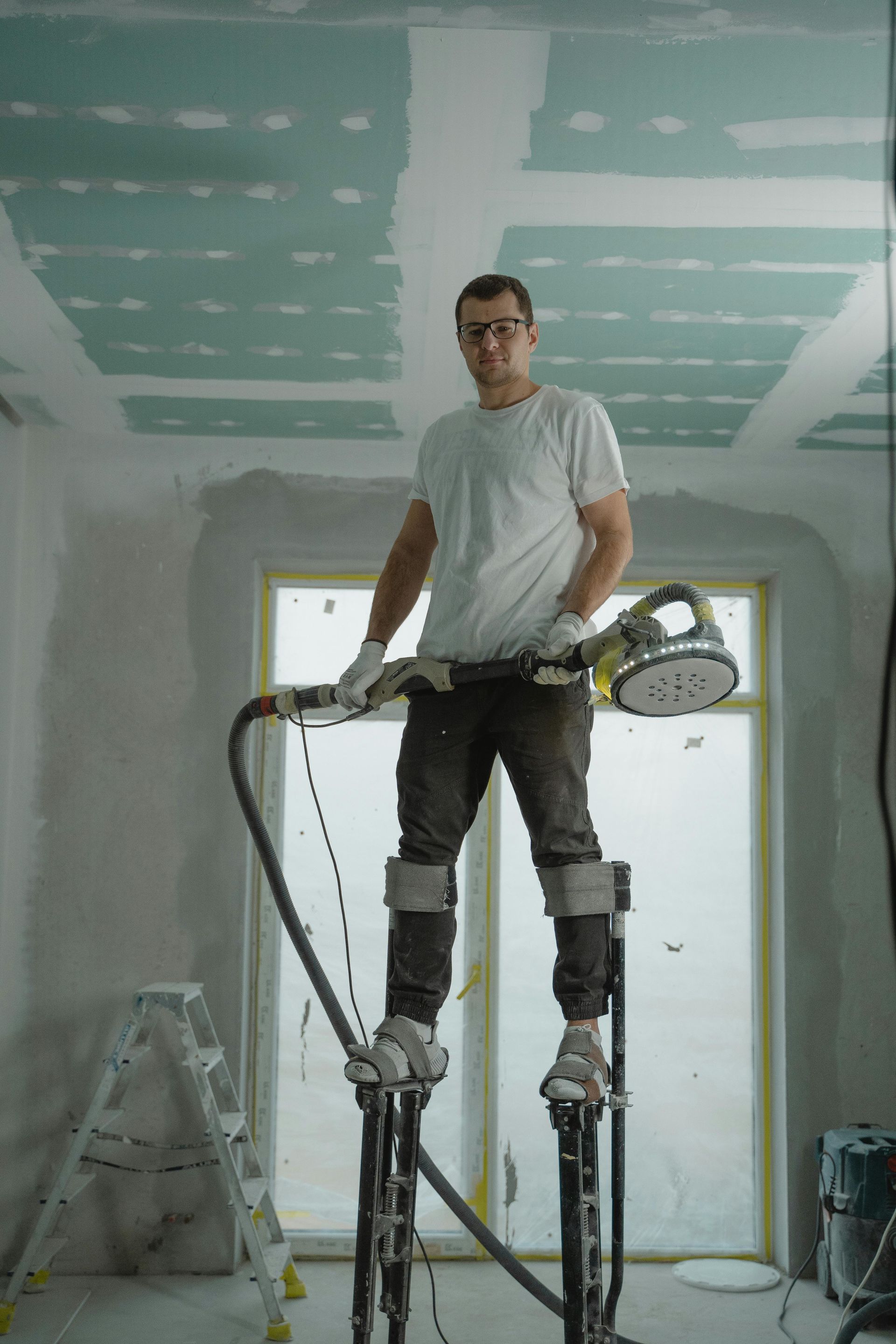 Man sanding drywall on stilts in a room under construction.