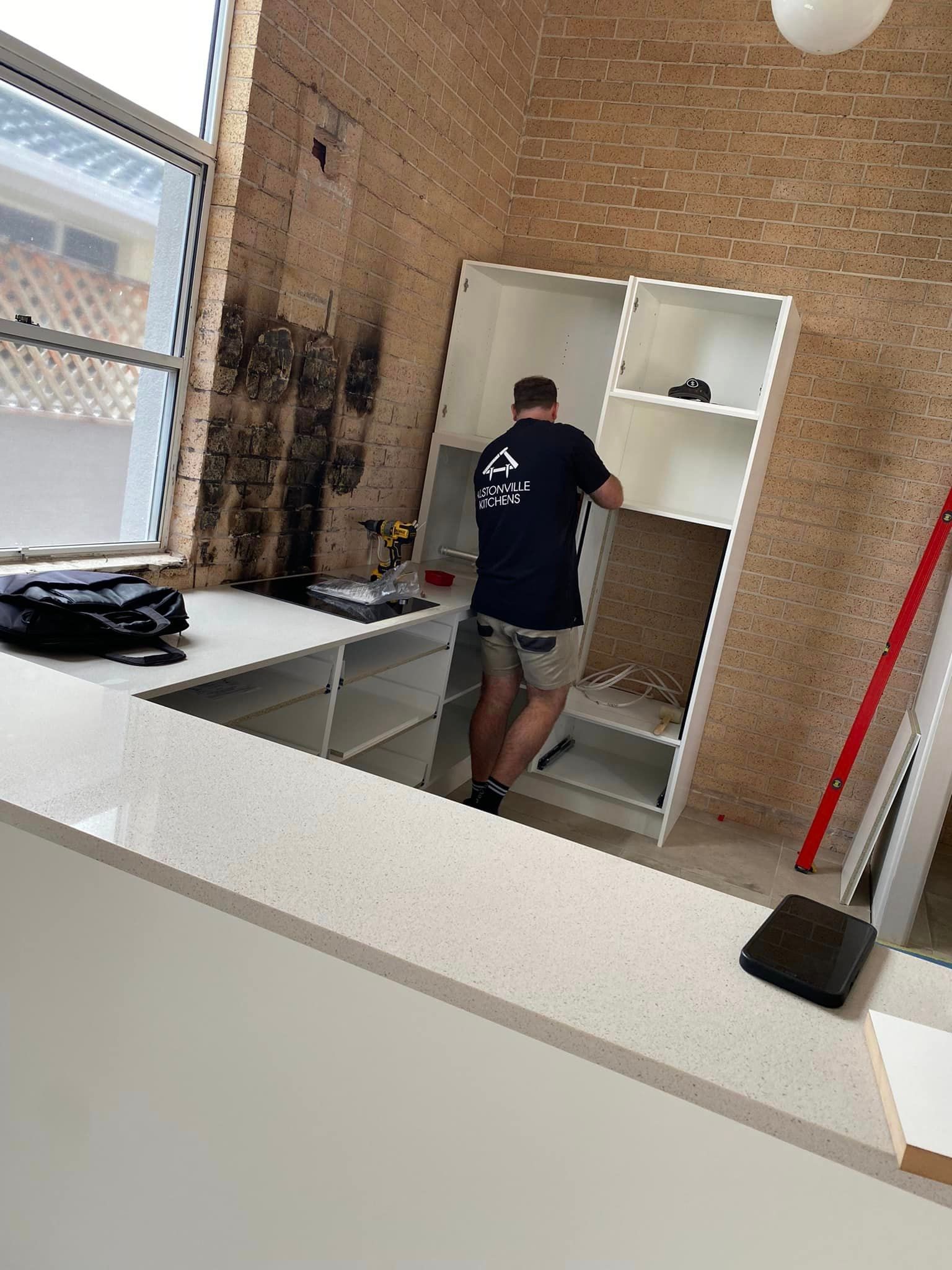 A Person Installing White Cabinets In A Kitchen Remodel. The Space Has Exposed Brick And A Window  — Jason Walsh Constructions in Ballina, NSW