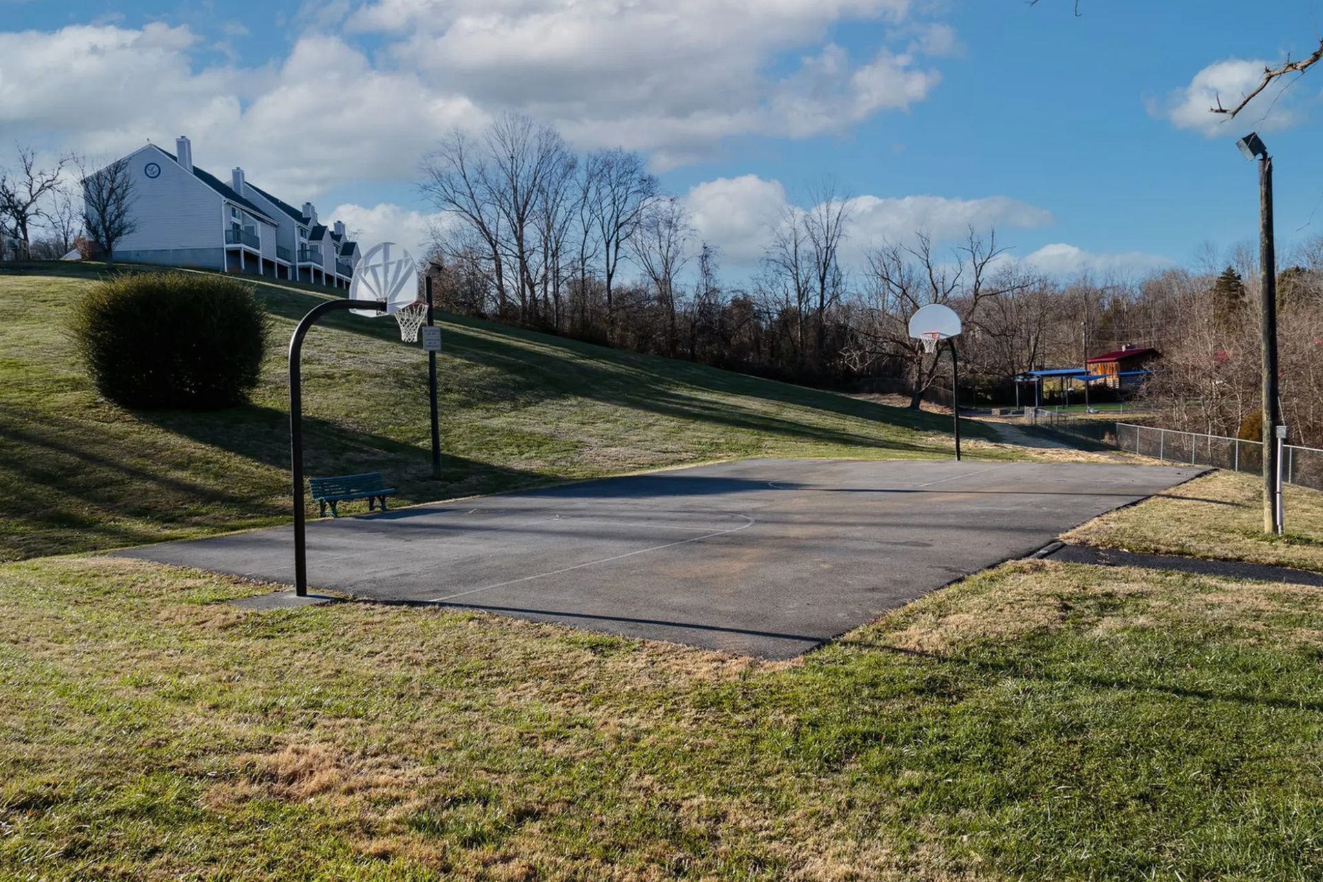 Basketball court on a grassy hill with two hoops. Buildings and trees in the background on a sunny day.