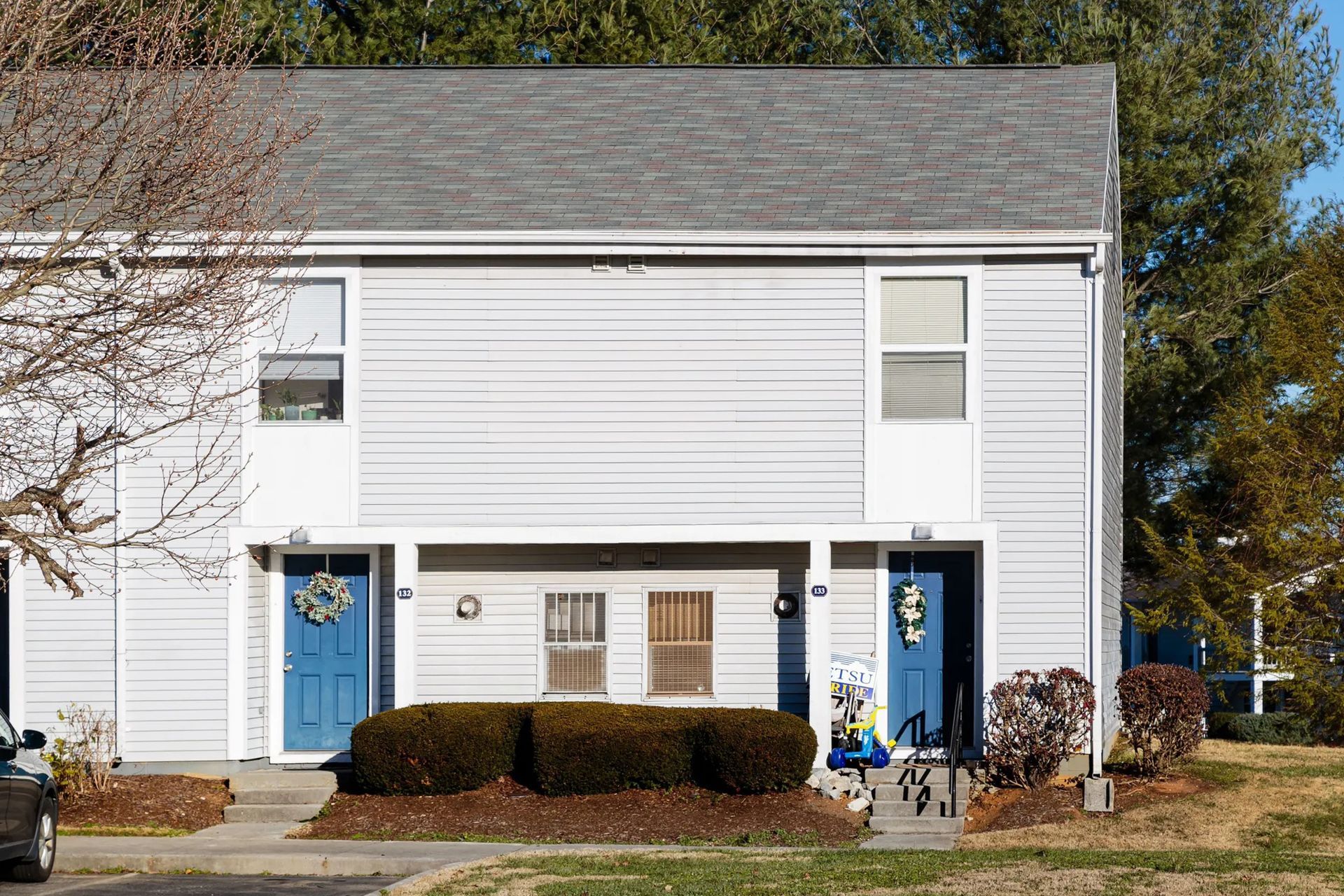 Two-story duplex with blue doors, light blue siding, and gray roof; bushes and bare trees in front.