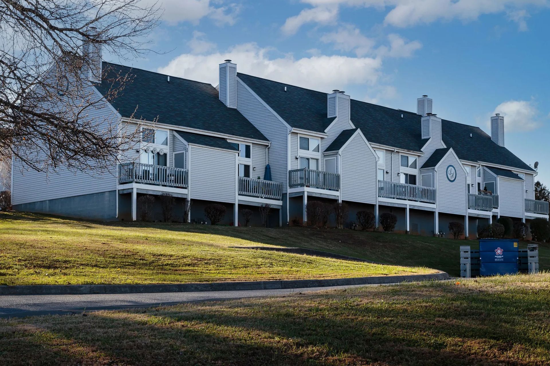 Row of townhouses with gray siding, balconies, and chimneys on a grassy hill under a blue sky.