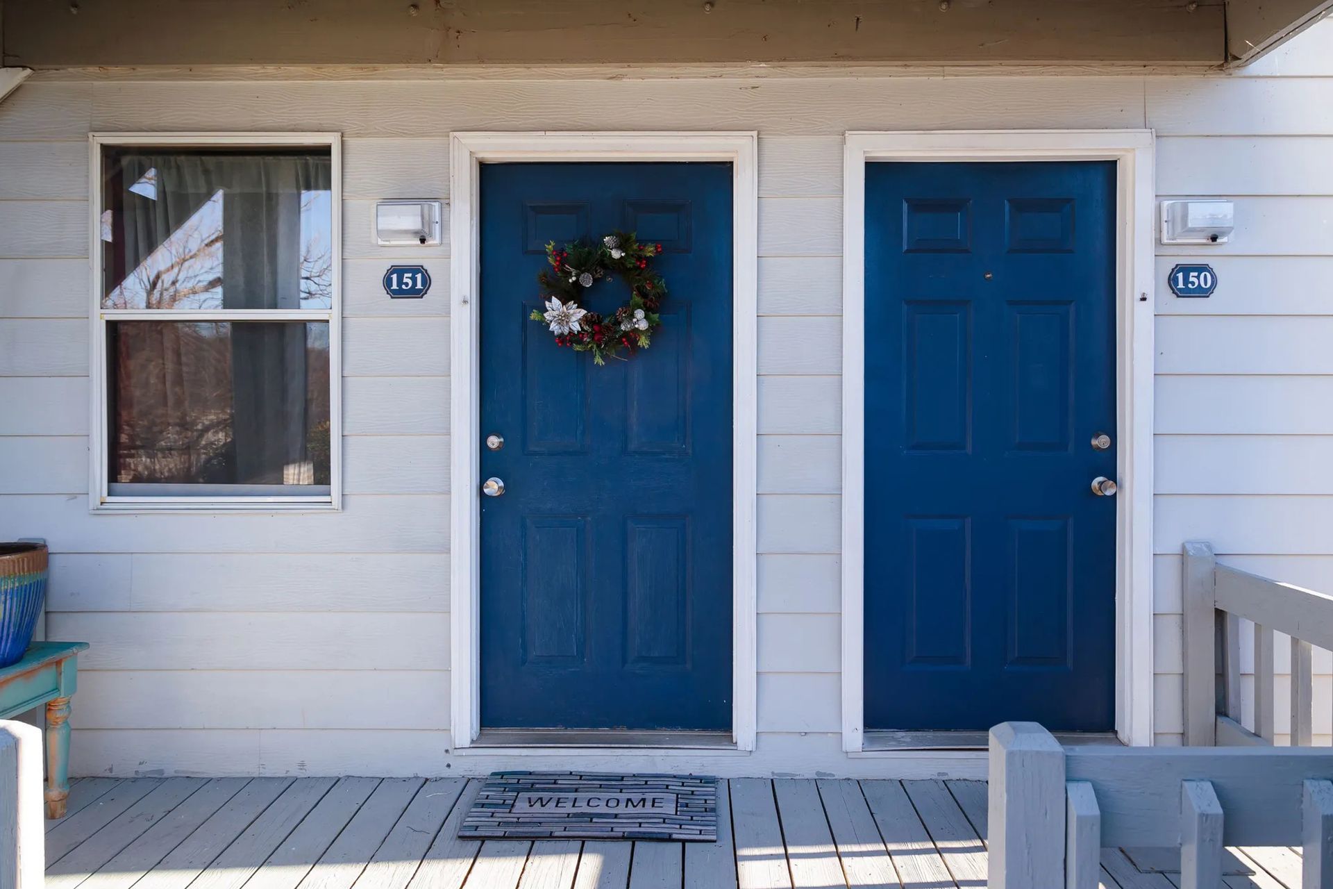 Exterior of two blue doors with white trim, between a window and railing, with a welcome mat.